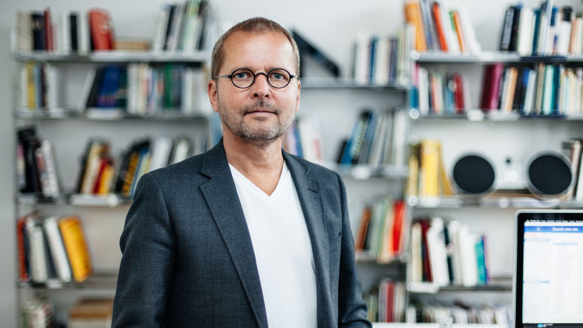 Florian is standing in front of the bookshelf in his office in a white T-shirt and dark jacket. He has a three-day beard and wears glasses with round lenses.