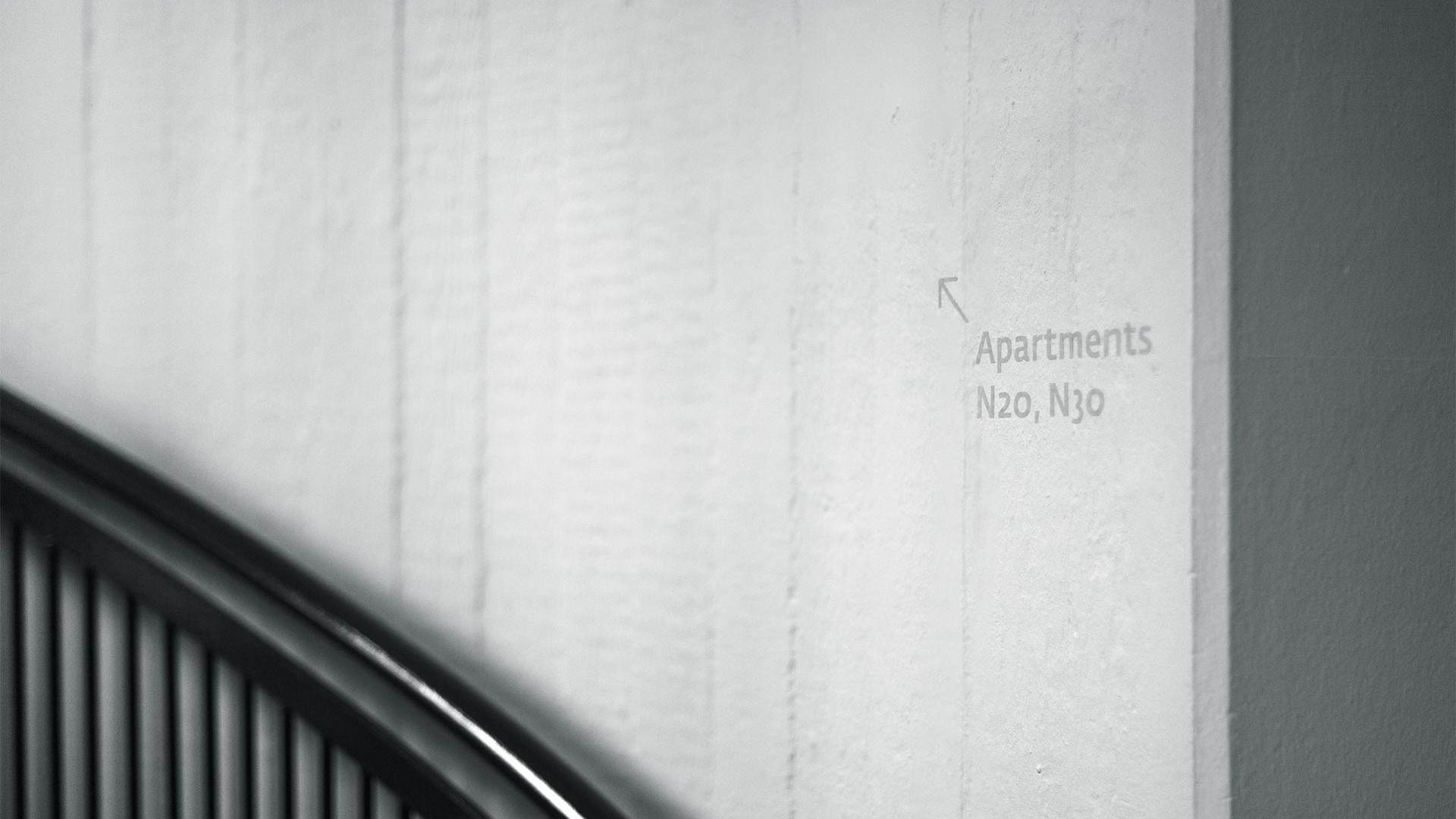 Auf einer rohen Betonwand mit einem schwarzen Treppengeländer verweisen dezente, hellgraue Buchstaben auf die Apartments im ersten Stock.